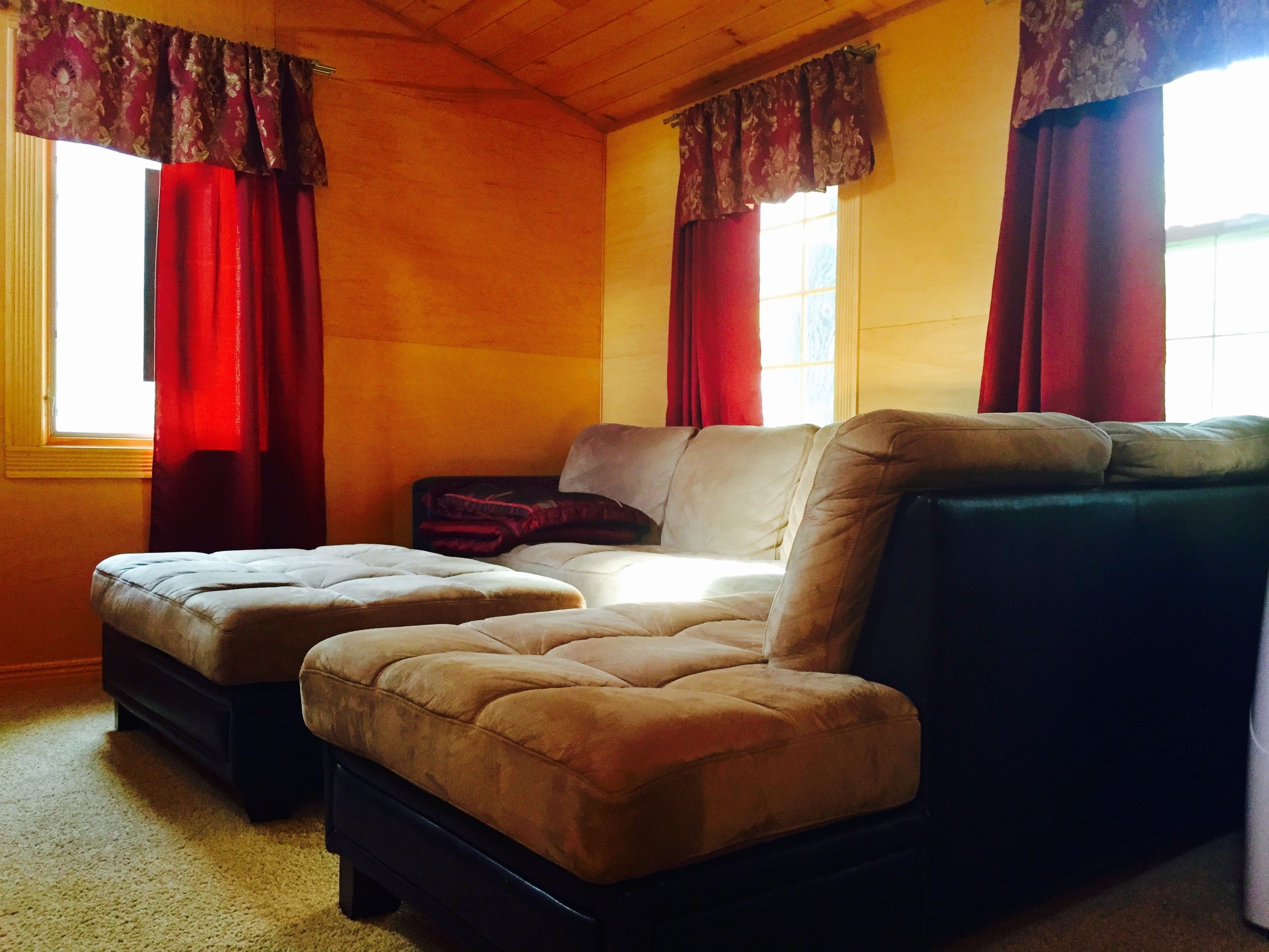 A cozy room with a sofa and ottoman set, wooden ceiling, and large windows with red curtains.