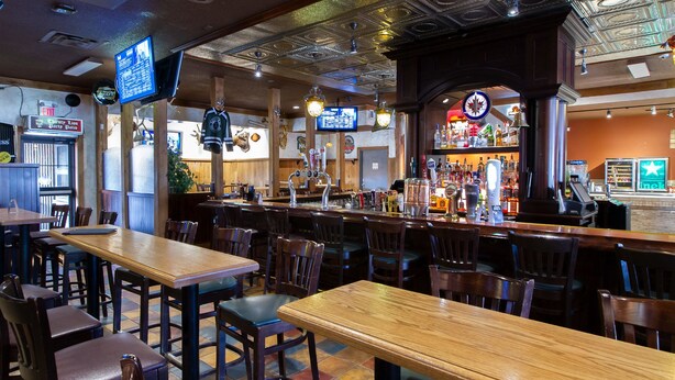 A bar area with wooden tables and chairs, a bar counter with various bottles and a cash register, and a television mounted on the wall. - Park West Inn (Winnipeg)