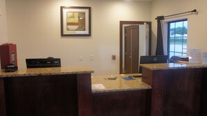 A reception area with a desk, a framed artwork on the wall, a fire alarm, and a window with a view of buildings. - Lux Inn and Suites (Three Rivers)