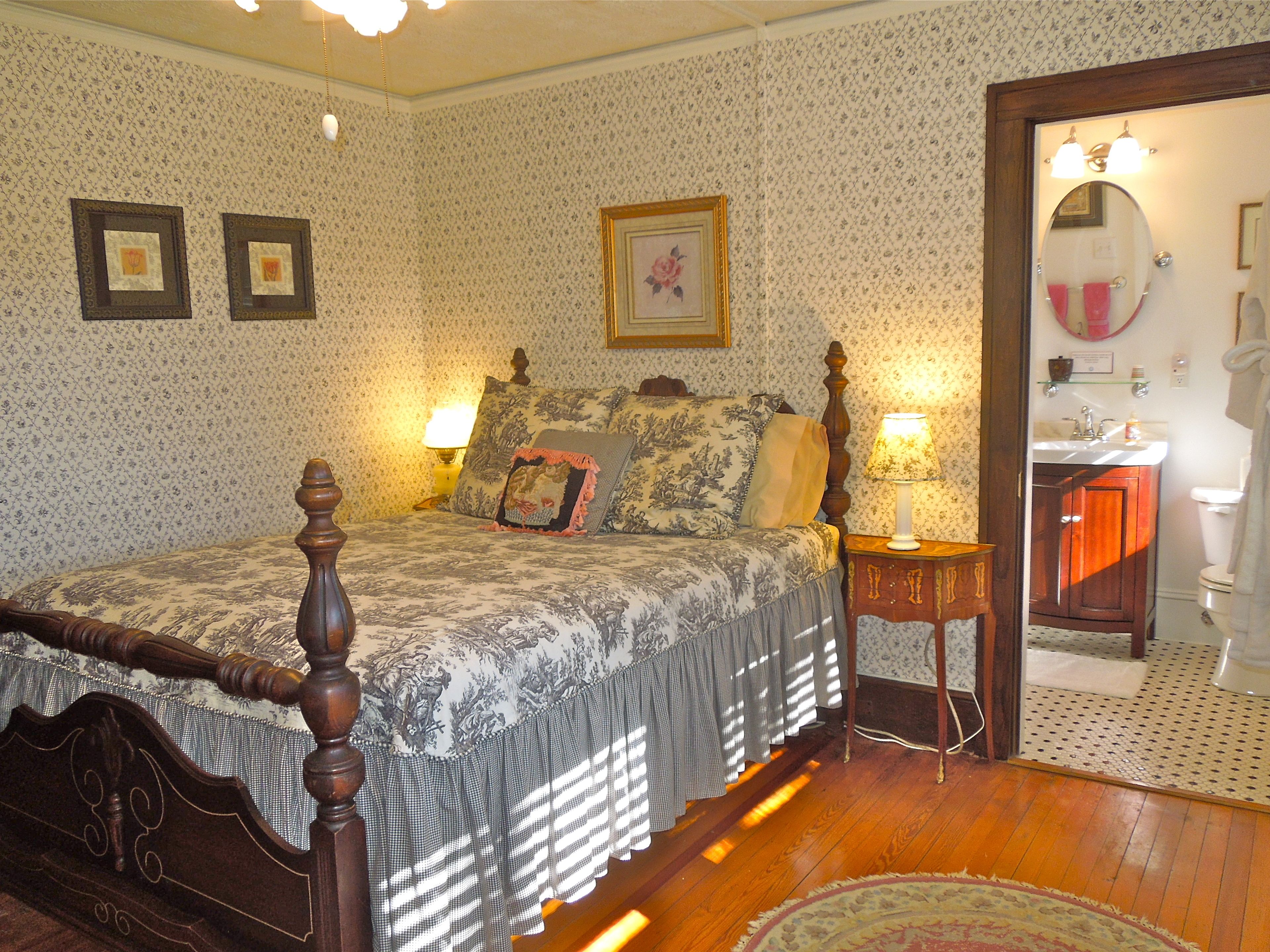 A bedroom with a four-poster bed, a nightstand with a lamp, and a bathroom visible through an open door.