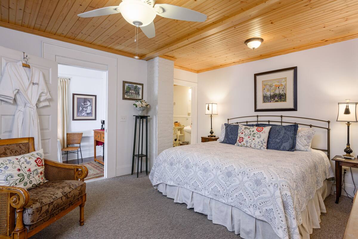 Une chambre avec un grand lit, un ventilateur de plafond en bois, une chaise, une table de chevet avec une lampe et un tableau encadré au mur.