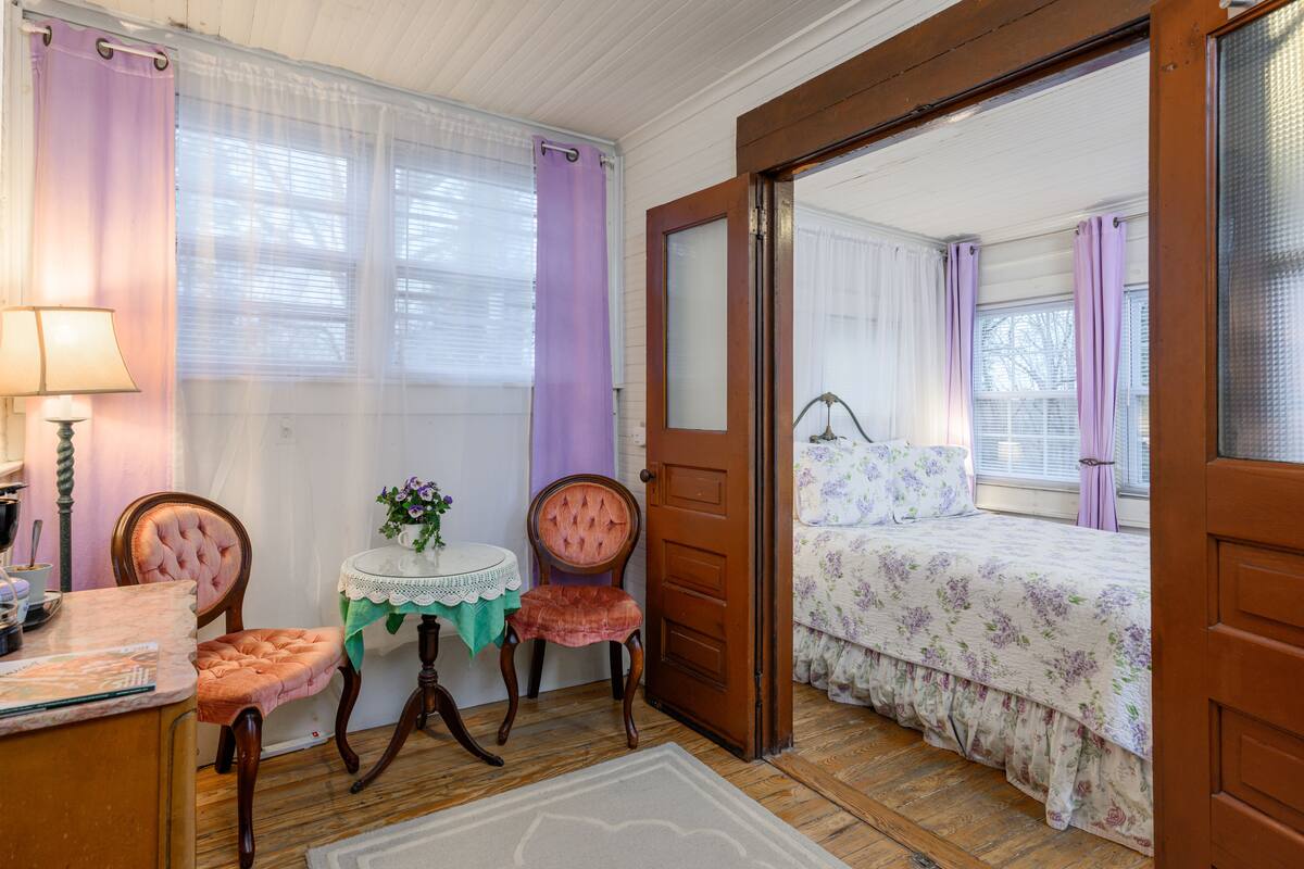 Une chambre avec une porte en bois, un lit recouvert d’une literie à motifs floraux, une petite table recouverte d’une nappe en dentelle et deux chaises dotées d’assises rembourrées.