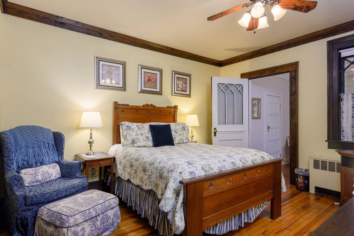 Une chambre avec un lit en bois, un fauteuil bleu, un pouf assorti et une table de chevet avec une lampe. Des tableaux encadrés ornent le mur, et une porte est agrémentée d’un panneau décoratif.