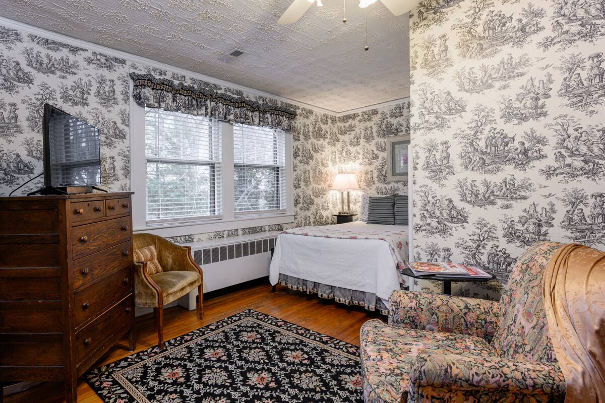 Une pièce comprenant une commode en bois, une chaise, un lit recouvert d’une couvre-lit à motifs floraux, une table de chevet avec une lampe et une fenêtre avec des stores.