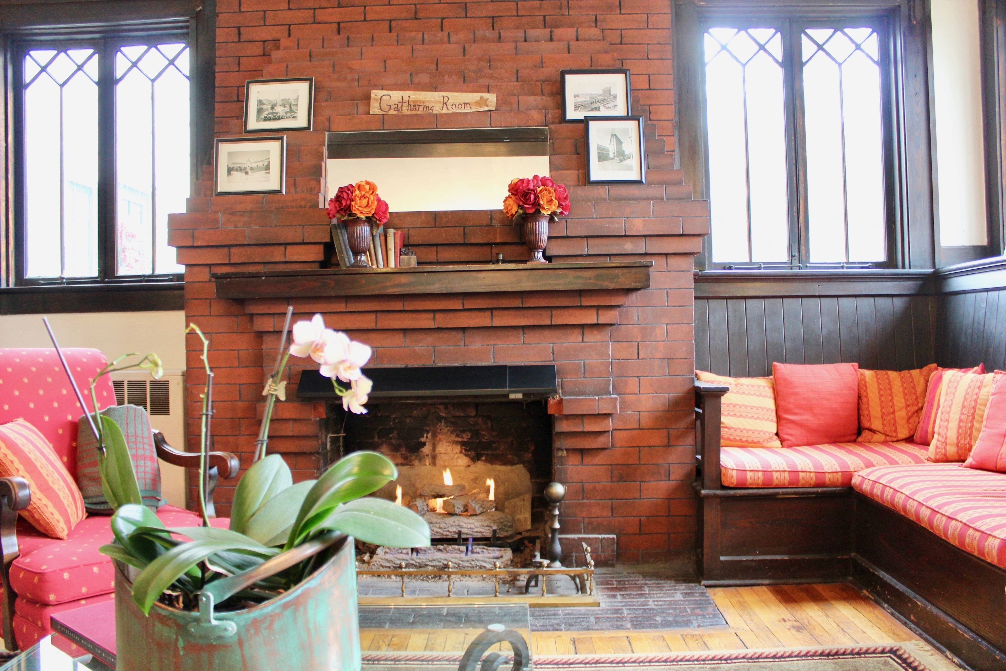 A cozy room with a brick fireplace, a sofa, and a chair. There are two vases with flowers on the mantel above the fireplace.