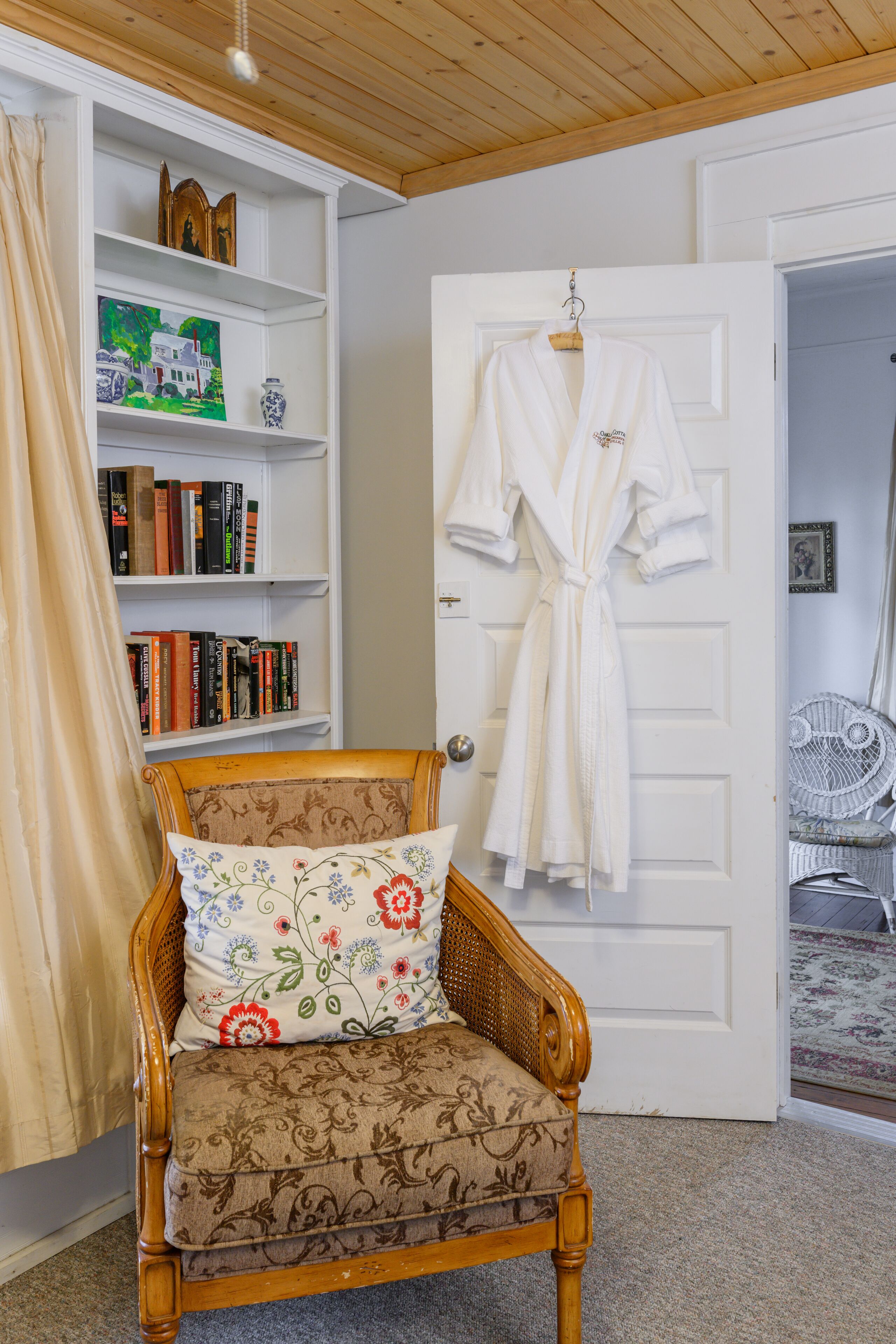 Una habitación con una silla de madera, una bata blanca colgada en la puerta y una estantería con libros.