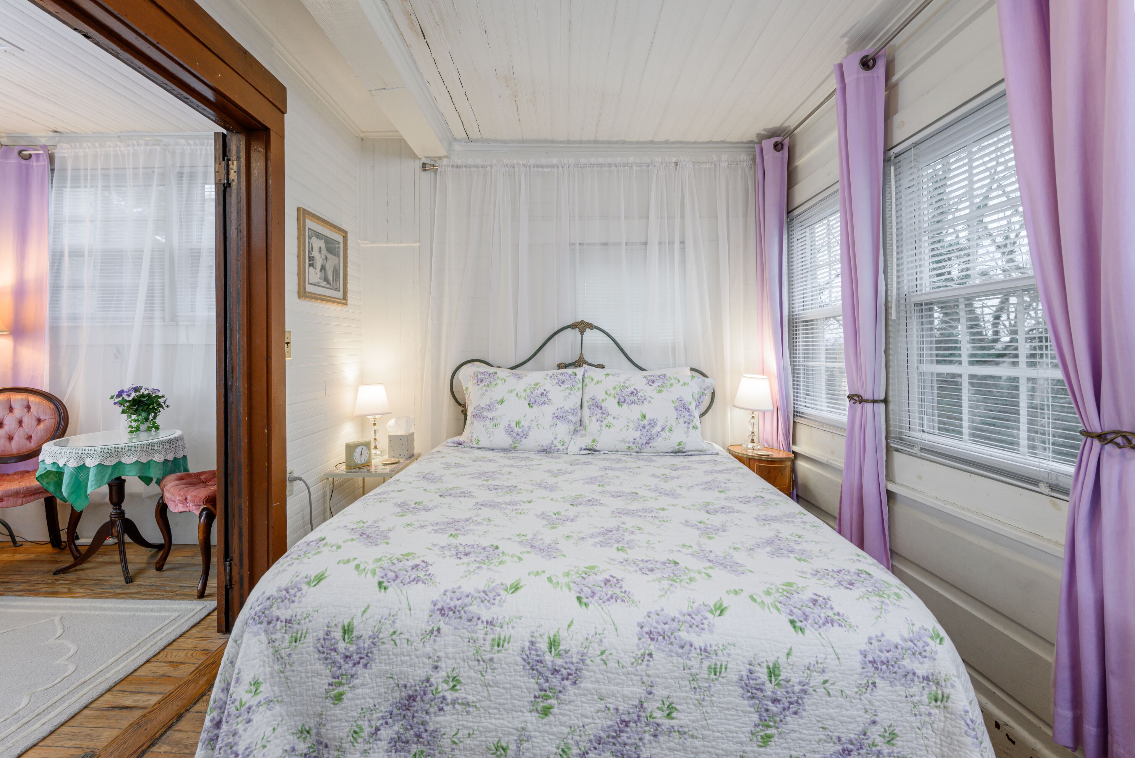 Un dormitorio con cubrecama floral, cabecera de madera, mesita de noche con lámpara y ventana con cortinas moradas.