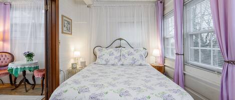 Un dormitorio con cubrecama floral, cabecera de madera, mesita de noche con lámpara y ventana con cortinas moradas.
