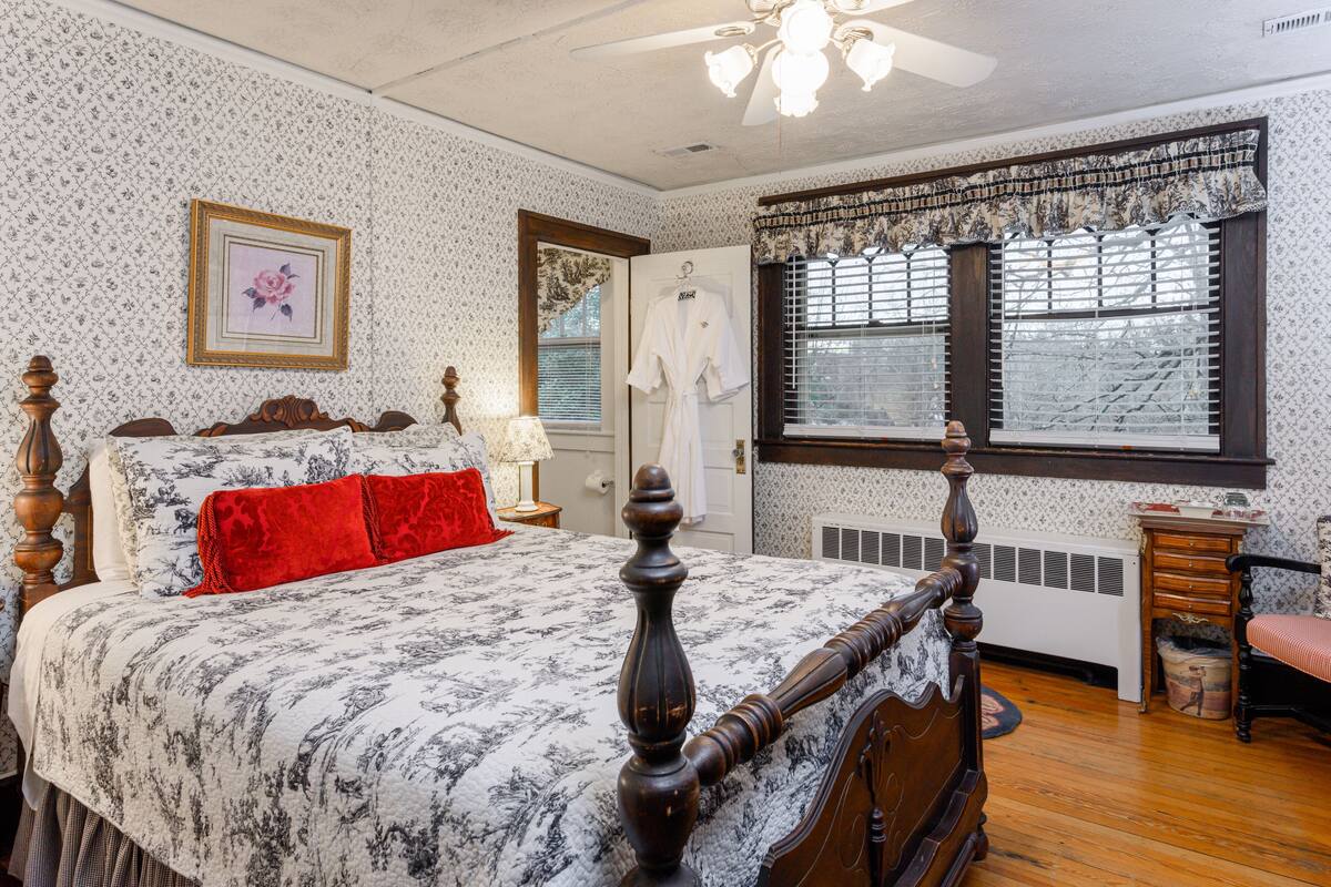 Une chambre à coucher avec un grand lit, une tête de lit en bois, une table de chevet avec une lampe, une fenêtre avec des stores et un radiateur.