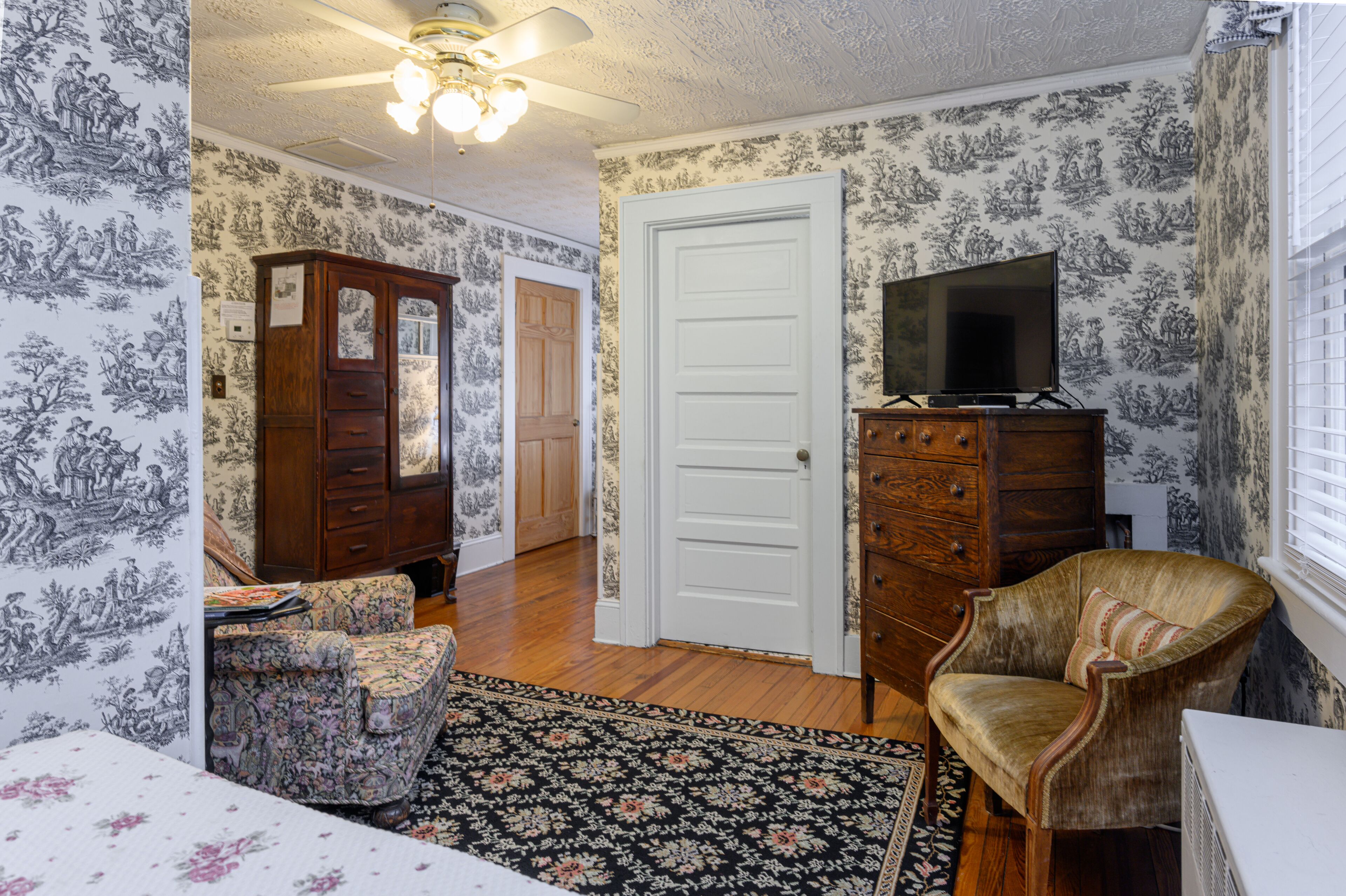A room with patterned wallpaper, a wooden dresser, a television on a stand, a floral rug, and a chair.