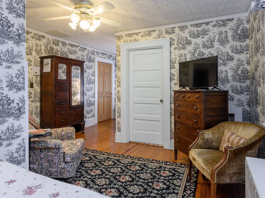 A room with patterned wallpaper, a wooden dresser, a television on a stand, a floral rug, and a chair.