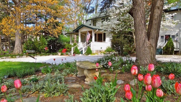 Une maison avec un porche et un drapeau, entourée d’un jardin où poussent des tulipes et où se trouve un abreuvoir à oiseaux en pierre.