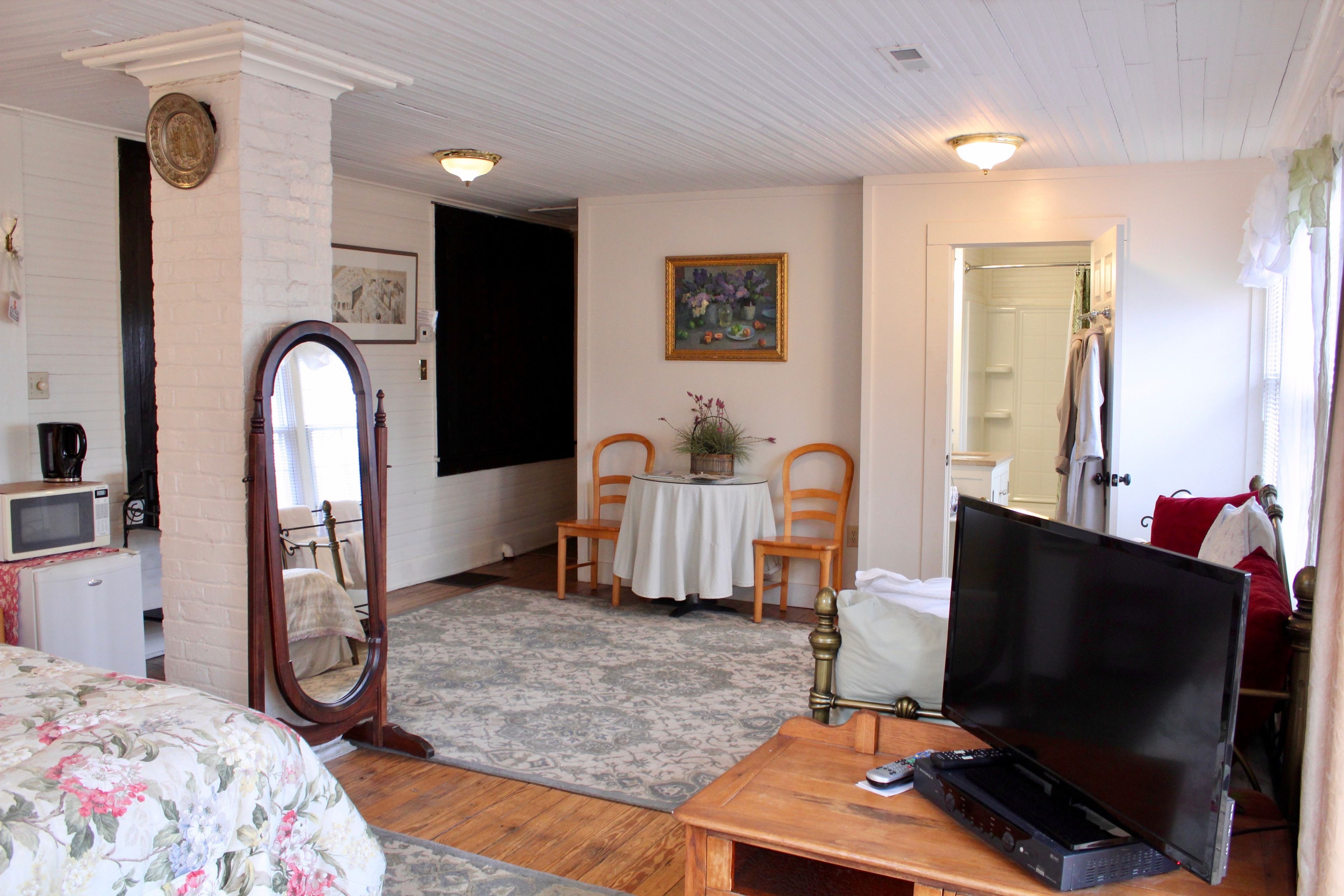 A room with a bed, a desk with a computer, a mirror, a small table with chairs, and a painting on the wall.