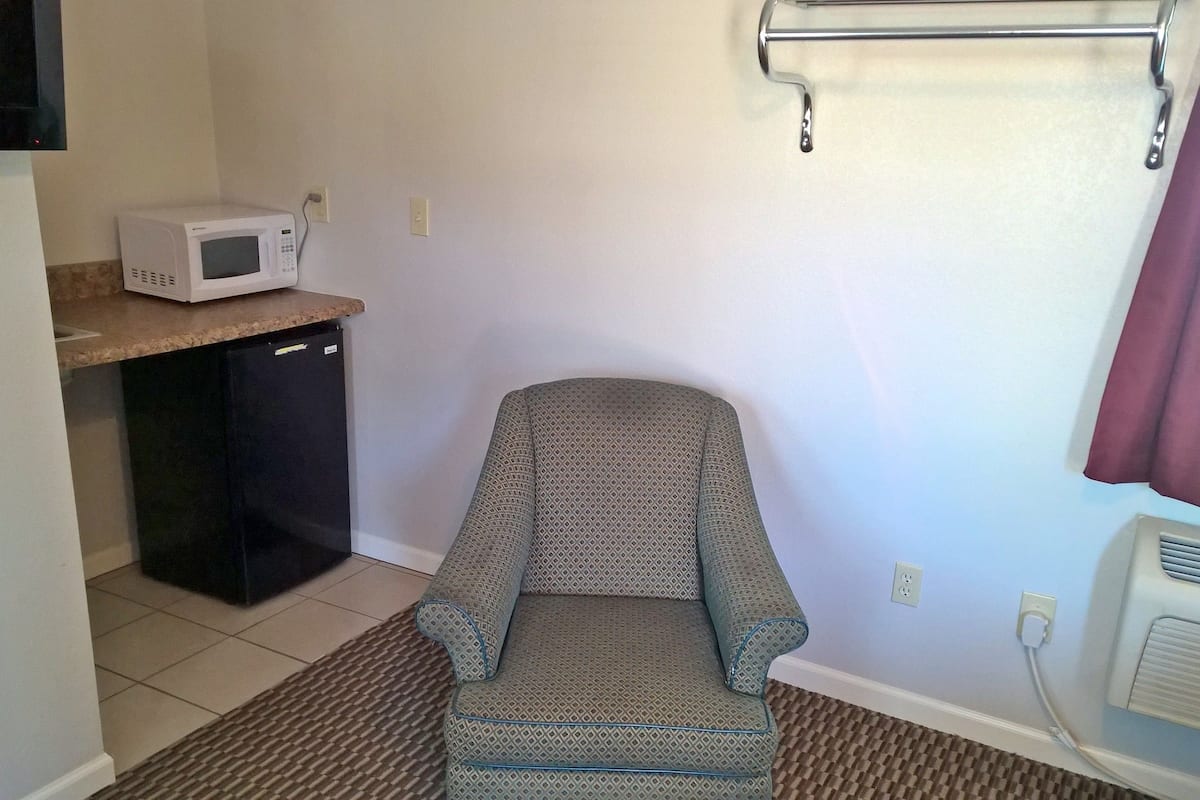 A compact room with a kitchenette, a small refrigerator, a microwave, a patterned chair, a radiator, and a curtain.