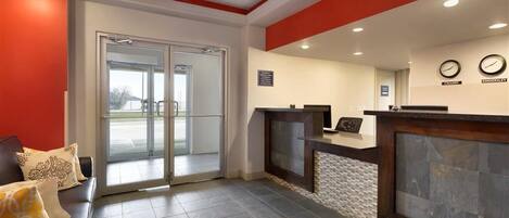 Un área de recepción con entrada por puerta de vidrio, un mostrador de recepción y un reloj en la pared.