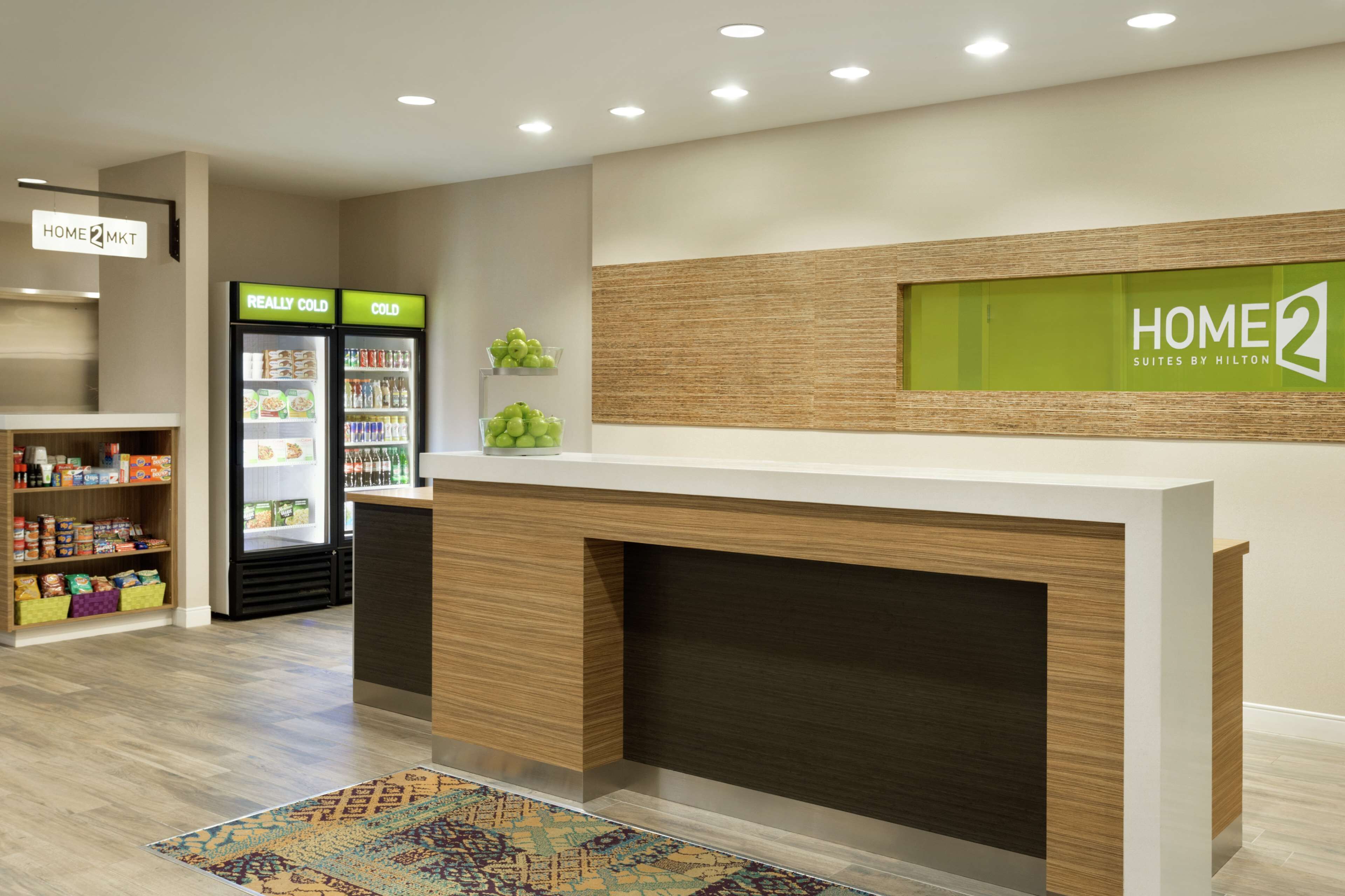 A hotel reception area with a reception desk, a 'Really Cold' and 'Cold' beverage refrigerator, a 'Home 2' sign, and a shelf with snacks.