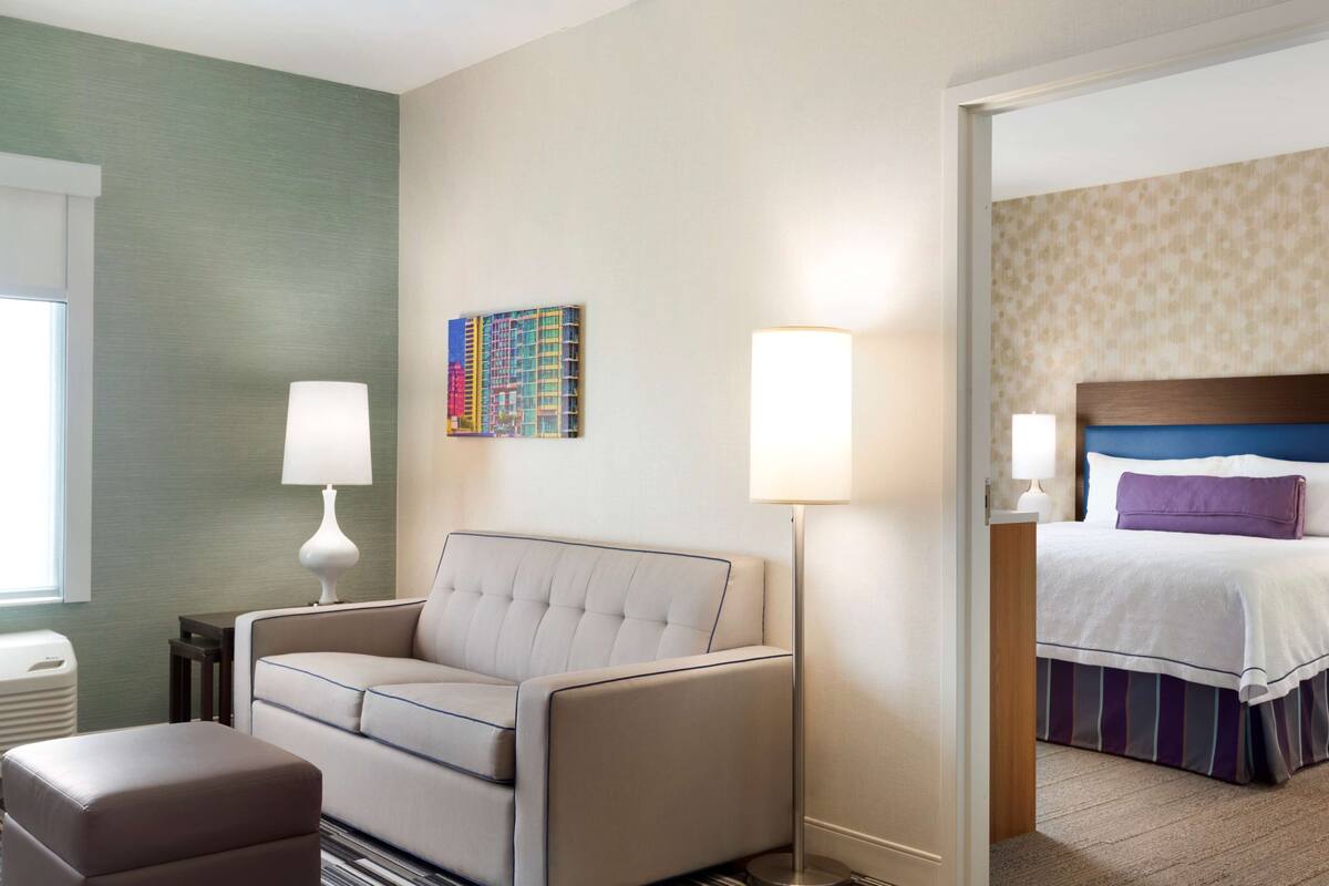 A hotel room with a sofa, a bed with a headboard, a lamp, a trash bin, and a wall with a colorful artwork.