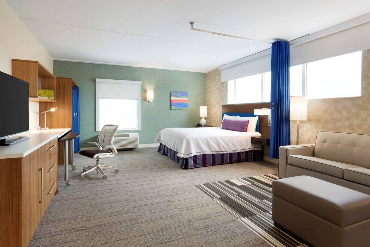 A hotel room with a bed, a desk with a flat-screen TV, a chair, a sofa, and a window with curtains.