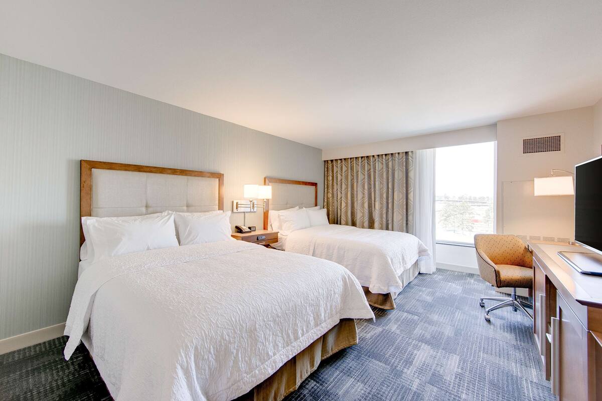 A hotel room with two beds, a desk with a chair, a television, and a window with curtains.