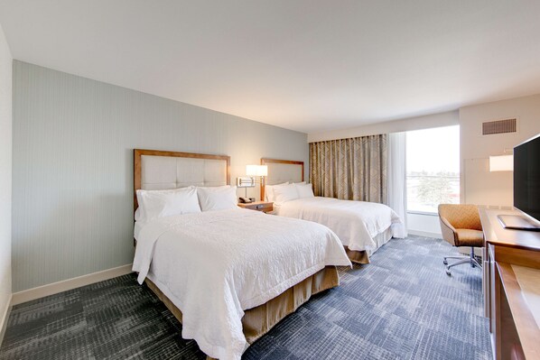 A hotel room with two beds, a desk, a chair, and a large window.