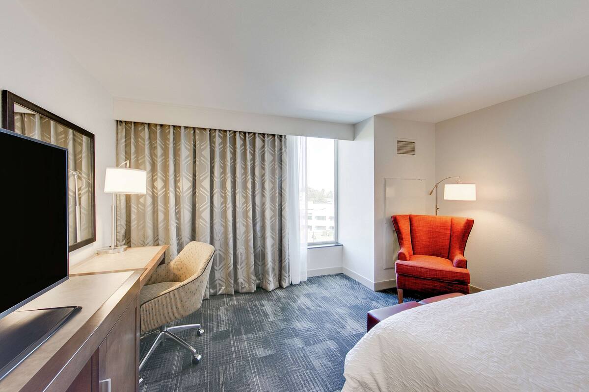 A hotel room with a bed, a desk with a TV, a chair, a red armchair, and a window with curtains.
