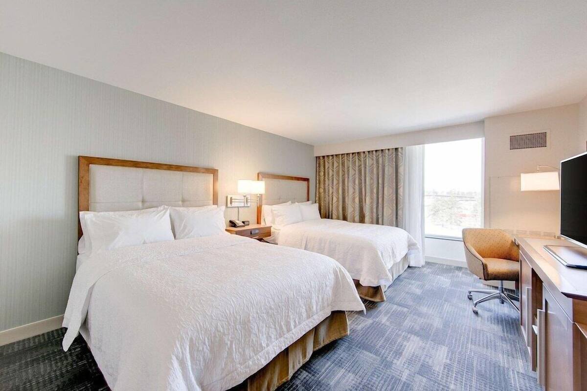 A hotel room with two beds, a desk, a chair, a TV, and a window with curtains.