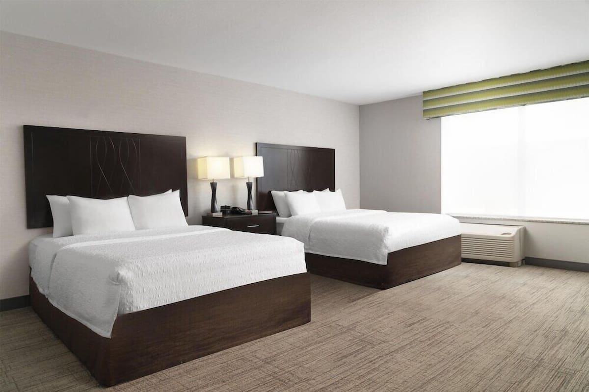 A hotel room with two beds, a nightstand with lamps, a window with blinds, and a wall-mounted air conditioner.