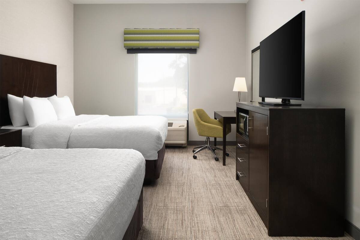 A hotel room with two beds, a desk, a television, and a window with blinds.