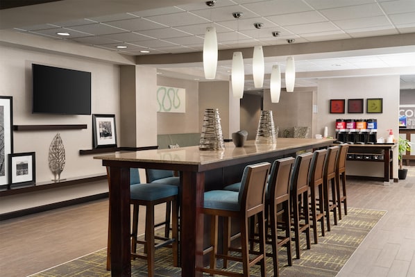 A hotel bar area with a long wooden bar, multiple bar stools, a flat-screen TV mounted on the wall, and a menu board displaying drink options.