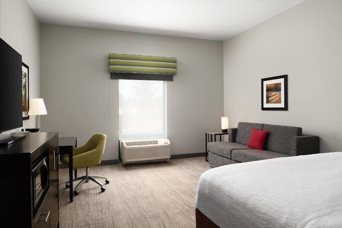 A hotel room with a bed, a desk with a chair, a sofa, a window with blinds, and a picture on the wall.