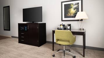 A hotel room with a dark wooden desk, a green office chair, a flat-screen TV, a framed picture on the wall, and a lamp.