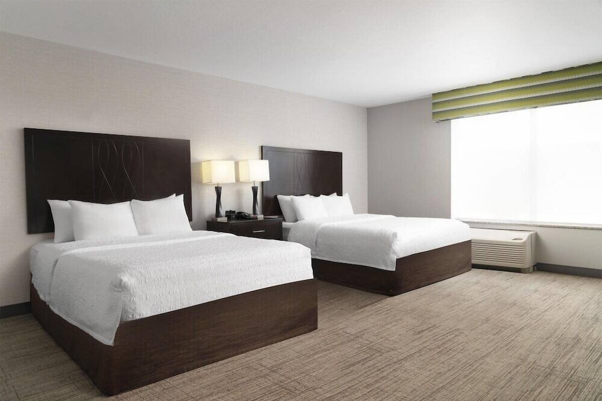 A hotel room with two beds, a nightstand with lamps, a window with blinds, and a wall-mounted air conditioner.