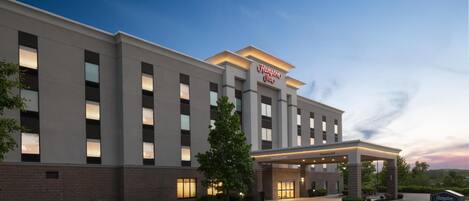 A modern hotel building with a prominent "Hampton Inn" sign, a covered entrance, and a landscaped driveway.