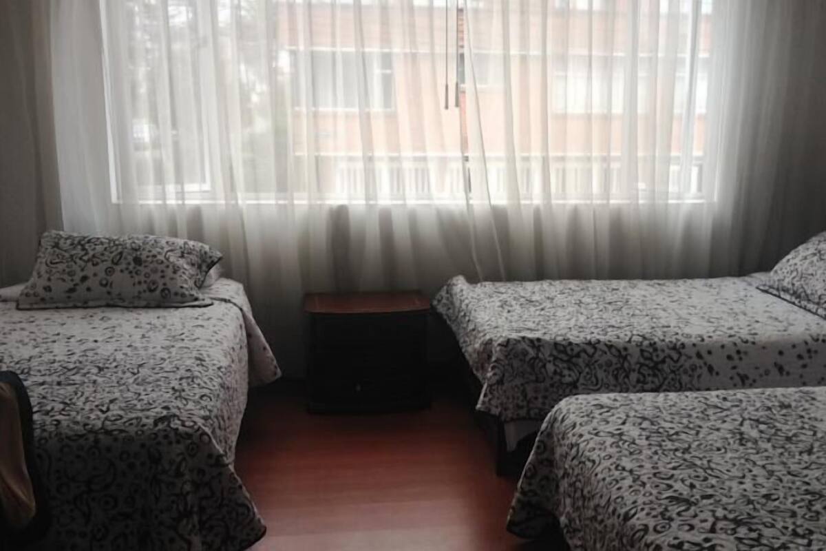 A room with two beds, each with patterned bedding, a small wooden stool, and sheer curtains.