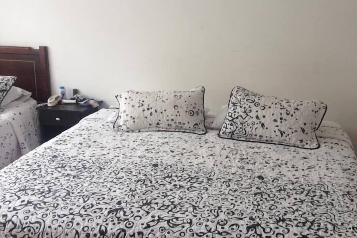 A bed with a patterned bedspread, two pillows with a similar design, a bedside table with a lamp, and a wall socket.