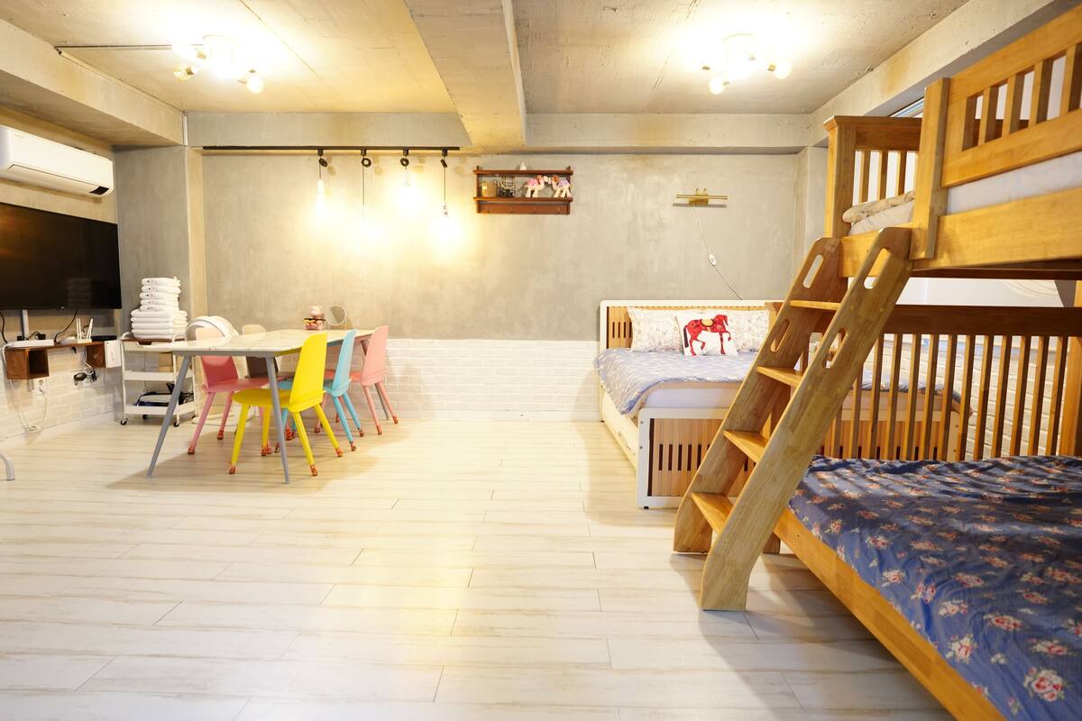 A room with a bunk bed, a desk with chairs, a television, and a wall-mounted shelf.