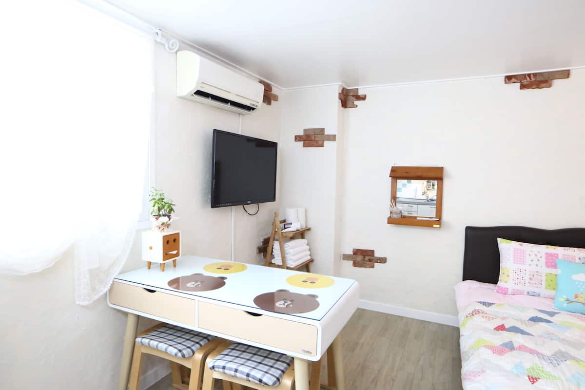 A bedroom with a bed, a TV mounted on the wall, a small table with chairs, and a window with a curtain.