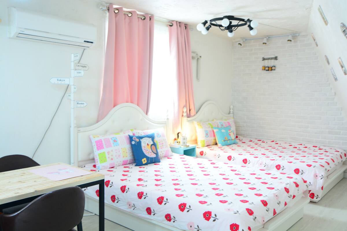 A bedroom with a bed, a desk, and a chair. There is a window with curtains, a wall-mounted air conditioner, and a light fixture.