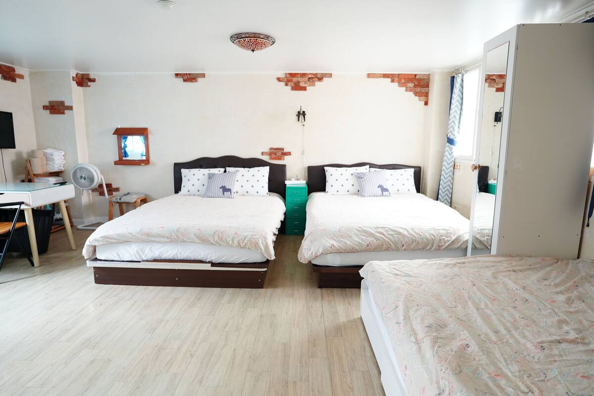 A double room with two beds, a desk, a chair, a fan, and a window with a view of the outside.