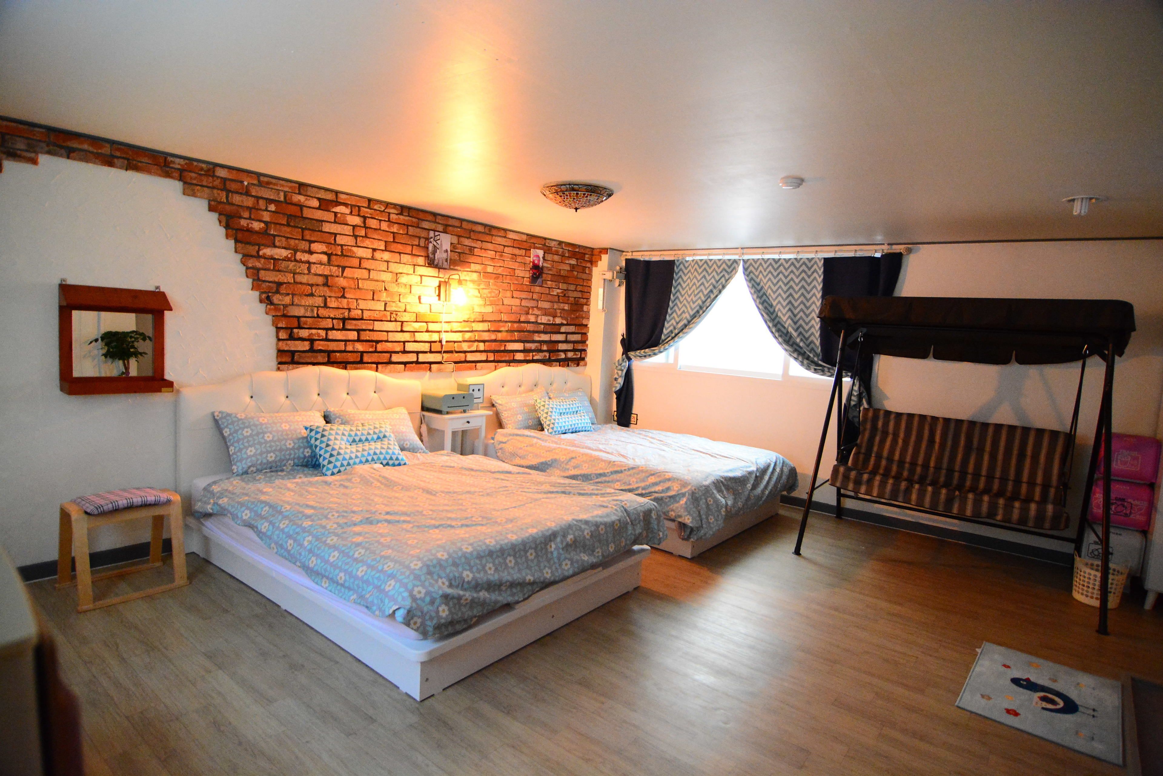 A bedroom with a brick wall, two beds with patterned bedding, a small bedside table, a bench, and a hanging swing.