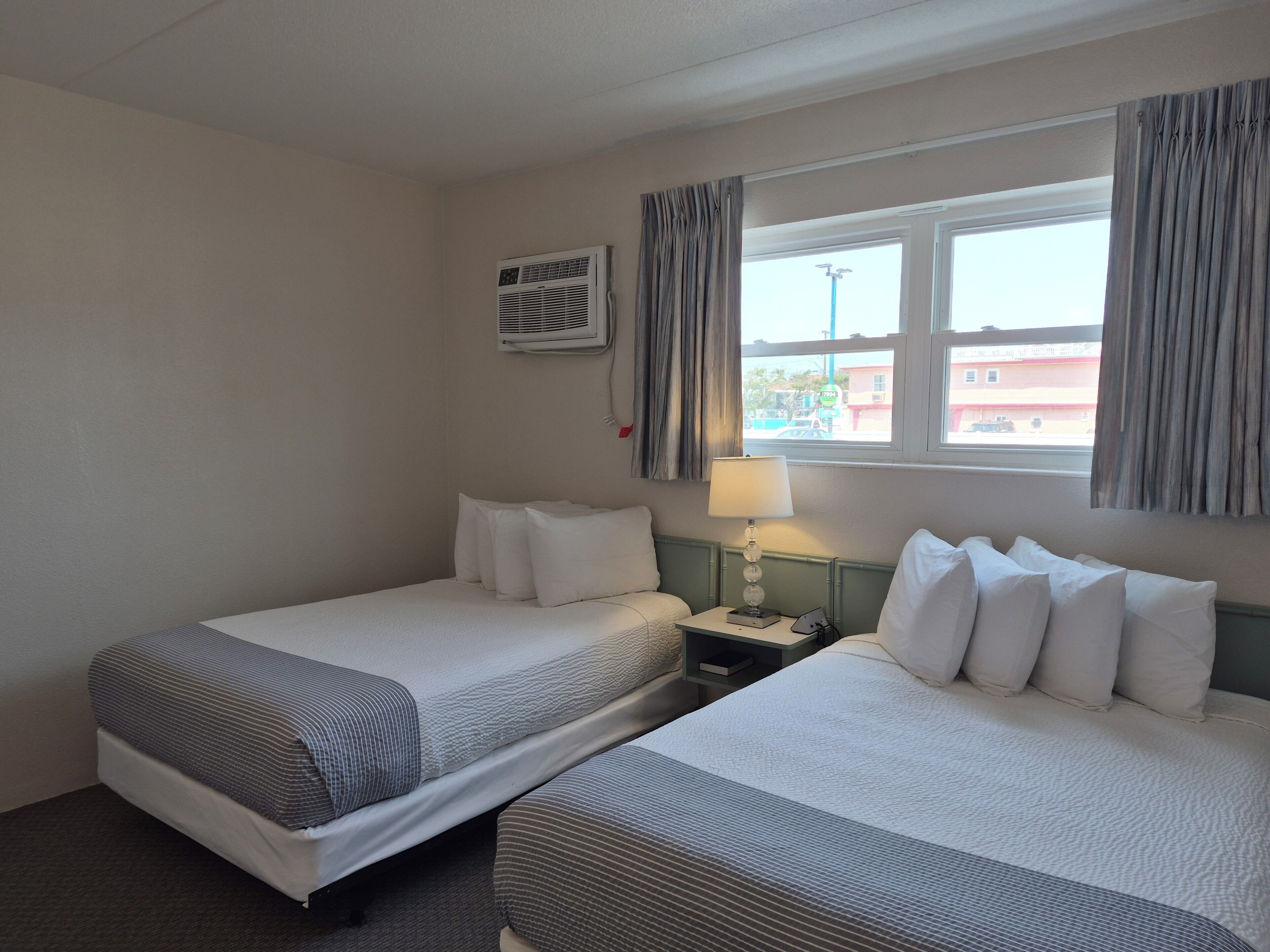 A hotel room with two beds, a nightstand, a lamp, an air conditioning unit, and a window with curtains.