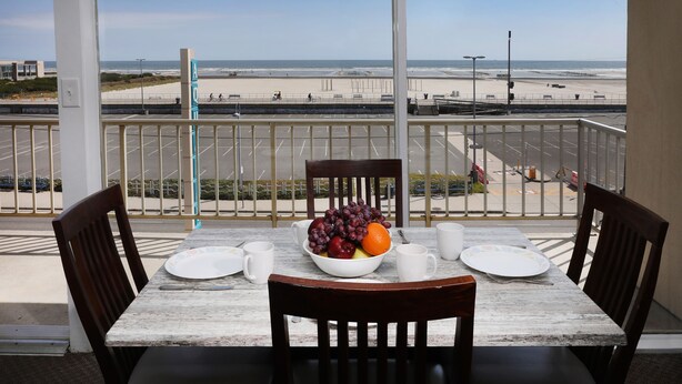 A dining table with chairs, a bowl of fruit, and white plates on a balcony overlooking a beach. - Aquarius Oceanfront Inn (Wildwood)
