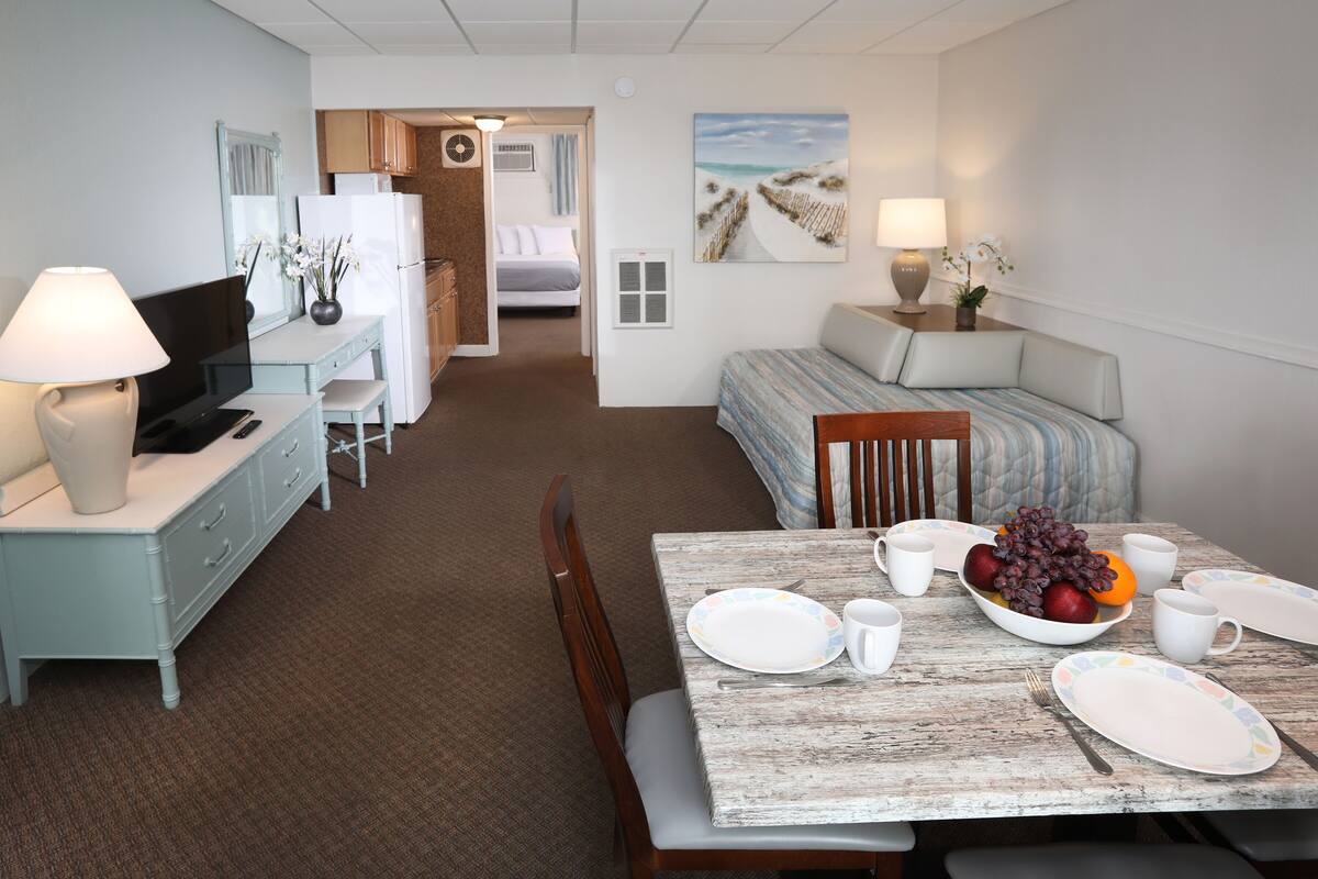 A hotel room with a dining table set for two, a bed with a view, a kitchenette, and a bathroom visible through an open door.