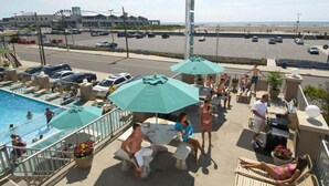 A rooftop pool area with lounge chairs, umbrellas, and a view of the beach and parking lot. - Aquarius Oceanfront Inn (Wildwood)