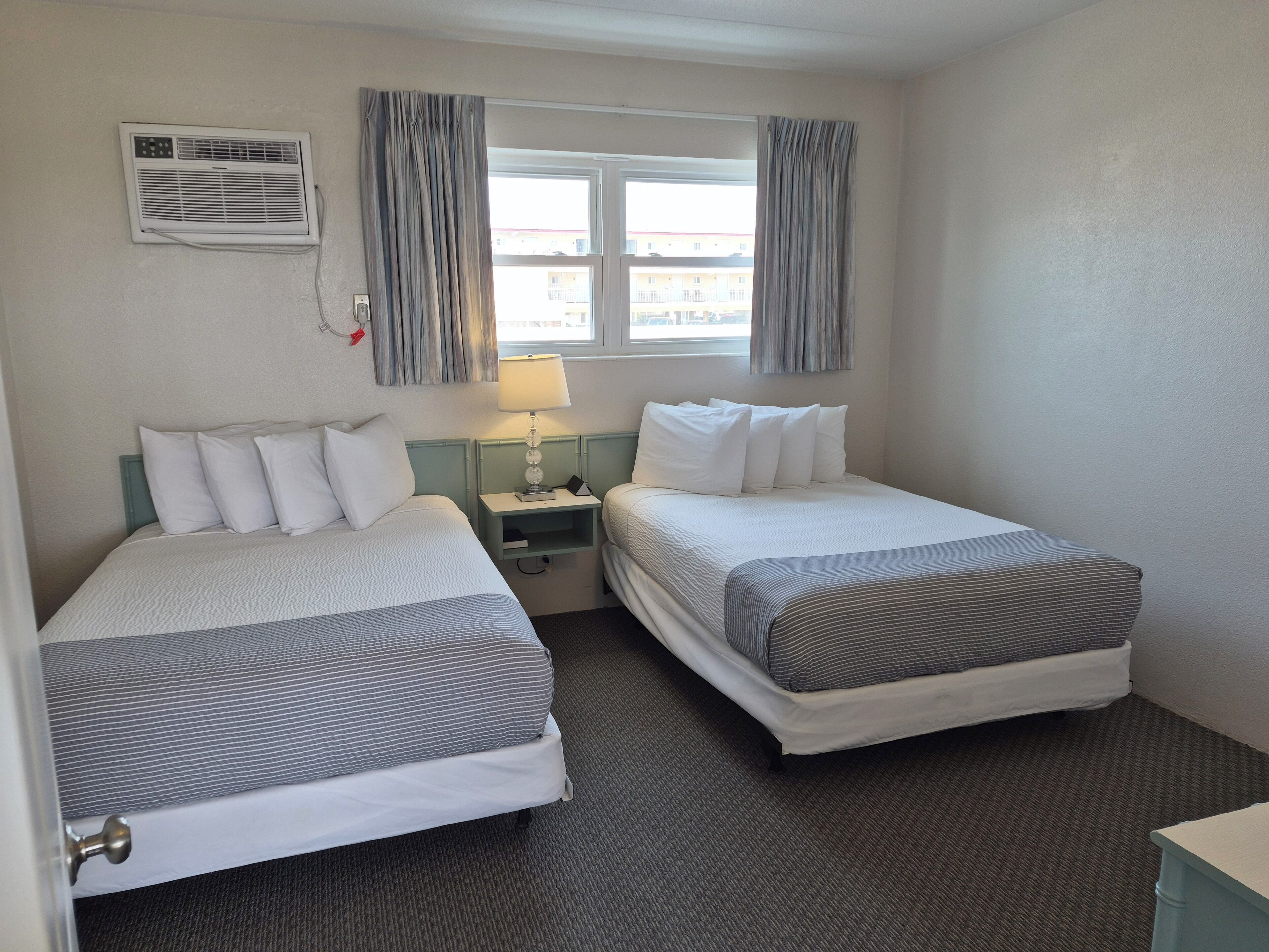 A hotel room with two beds, a window with curtains, a bedside table with a lamp, and an air conditioning unit.