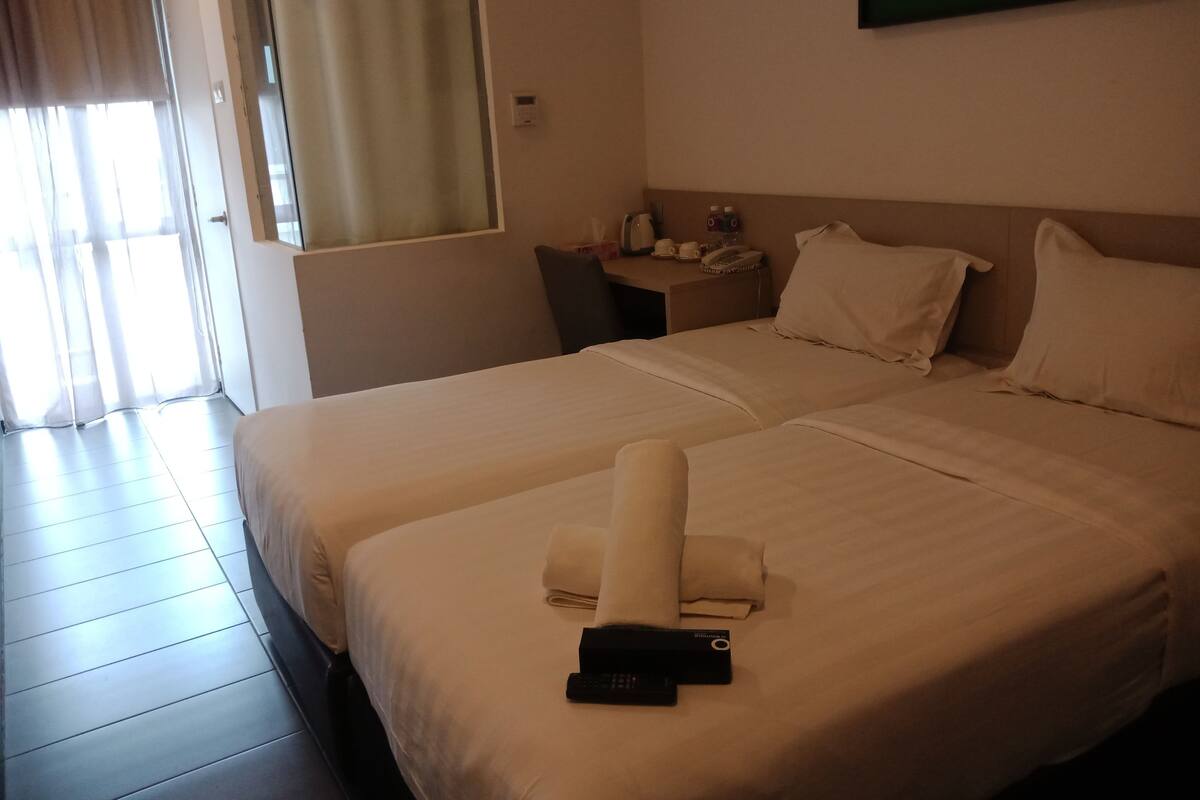 A hotel room with two beds, a desk with a phone and a kettle, and a window with curtains.