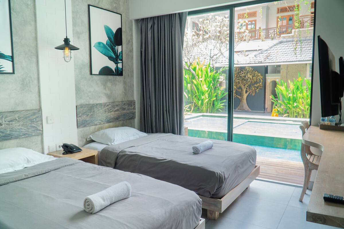A hotel room with two beds, a wooden desk, a telephone, and a large window with a view of a garden and building.