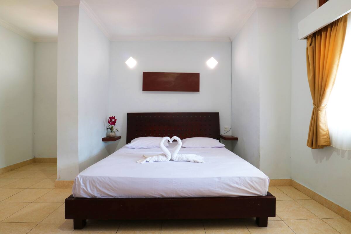 A neatly made bed with a heart-shaped towel arrangement, a wooden headboard, a small bedside table with a vase of flowers, and a wall-mounted light fixture.