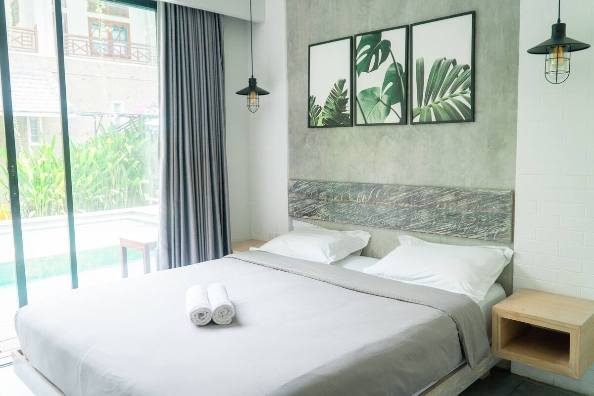 A hotel room with a bed, a nightstand, a window with curtains, and a view of greenery outside.
