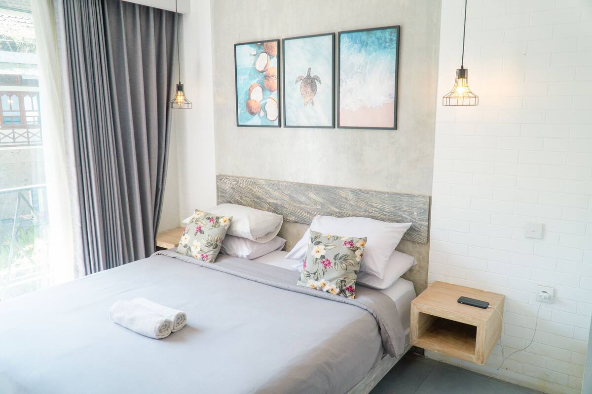 A neatly made bed with a grey bedspread and floral pillows, a wooden nightstand with a remote control, and a wall with three framed pictures depicting tropical scenes.